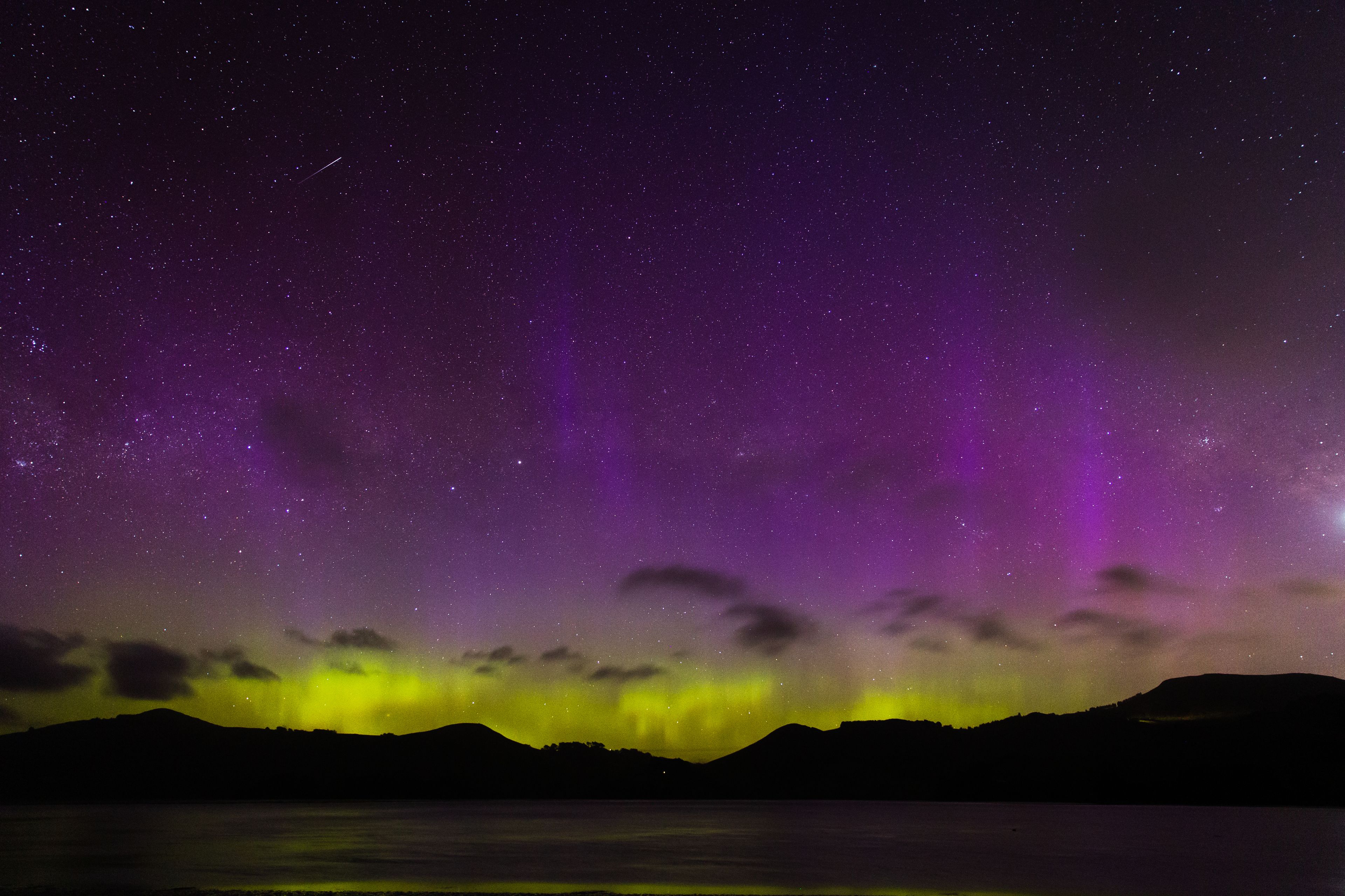 An aurora over a silhouette hillside showing a bright purple and yellow-green glow in the sky. There are stars visible and some clouds.