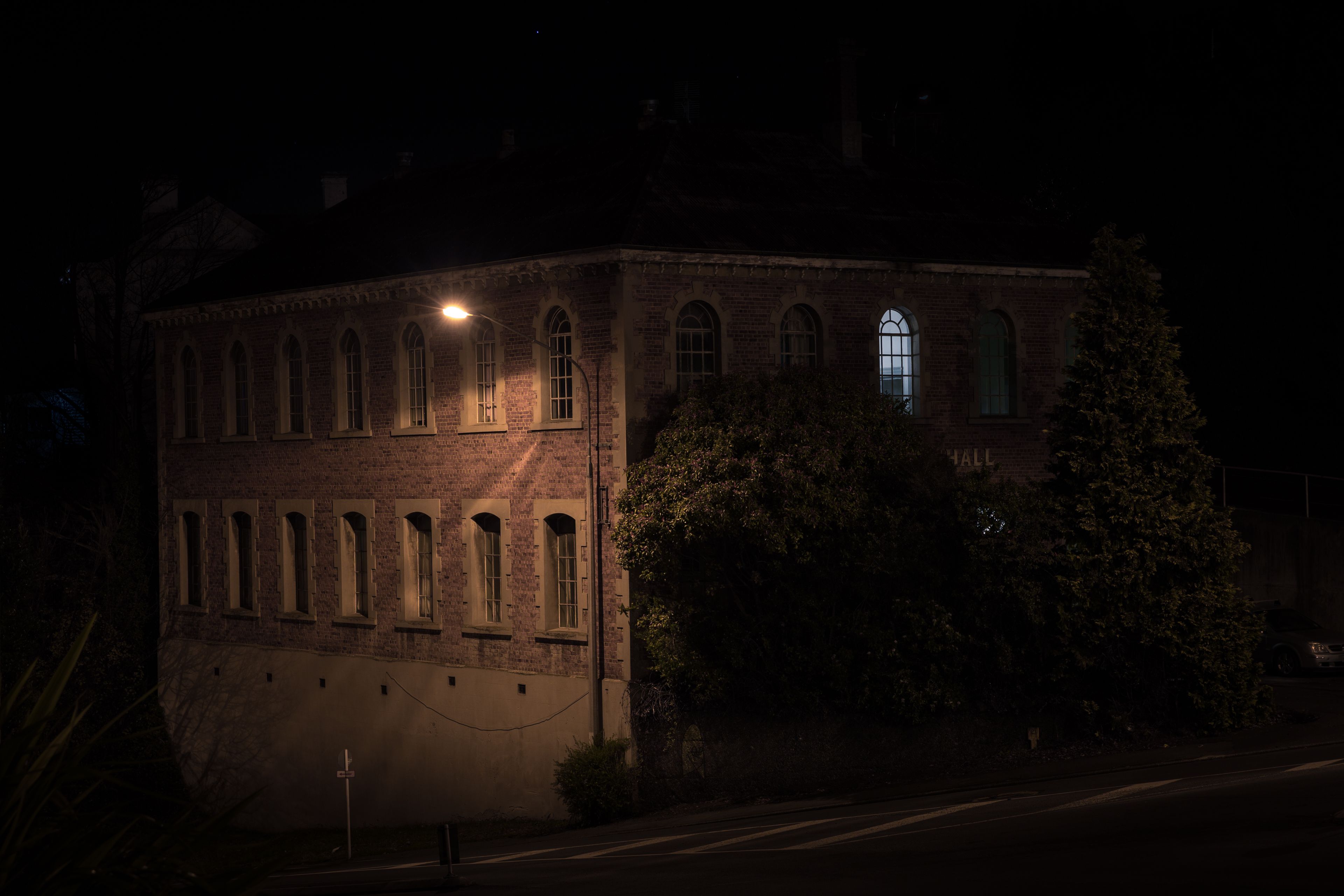 Two story brick building with one side dimly lit by a street light. There are trees growing against the building on the unlit side with a single window on the top floor in the middle glowing from a light inside.